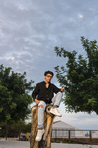 Young transgender man with glasses posing on a log outdoors.