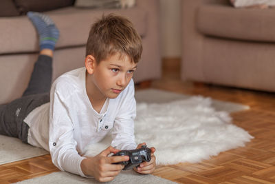Boy holding video game remote control while lying down on floor in living room at home 