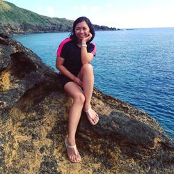 Portrait of smiling young woman sitting on rock by sea