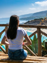 Rear view of woman sitting on wood against seashore and sky