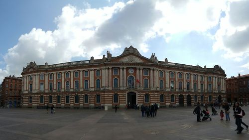 People walking in front of building against cloudy sky