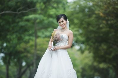 Portrait of smiling bride holding bouquet against trees