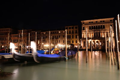 Boats moored in illuminated canal by buildings in city at night