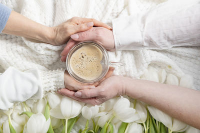 Four hands hold one of heart-shaped cofee cup, white tulips