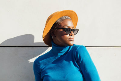 Serious african american female in stylish clothes and sunglasses standing near gray wall in sunny day