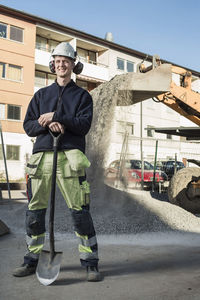 Full length portrait of smiling construction worker with shovel standing at site