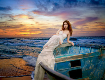 Portrait of young woman standing on beach against sky during sunset