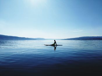 Side view of silhouette woman sitting on surfboard in sea against sky during sunny day