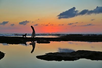 Silhouette woman with dog on rock at beach against sky during sunset