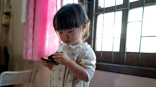 Girl holding object while standing against window at home