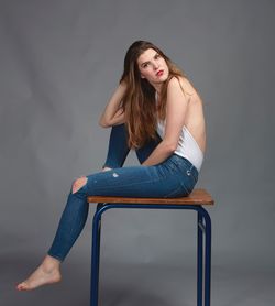 Side view full length portrait of young woman sitting on table against gray background