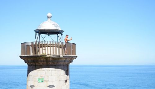 Lighthouse by sea against clear blue sky