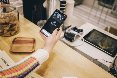 High angle view of woman holding mobile phone over credit card reader on table