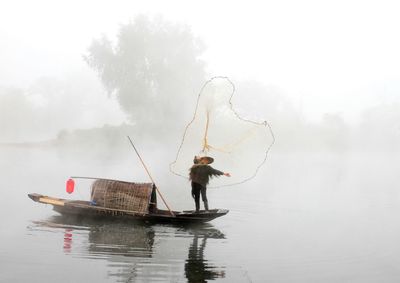 Man standing on boat while fishing in lake against sky