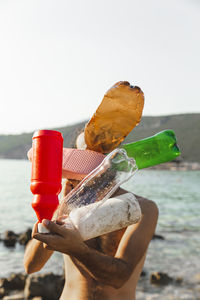 Young man holding empty plastic bottles and slipper in front of face at beach