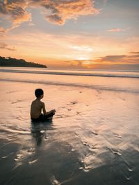 Boy sitting in sea against sky during sunset