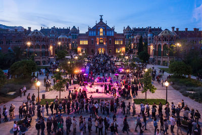 People on illuminated street amidst buildings in city against sky