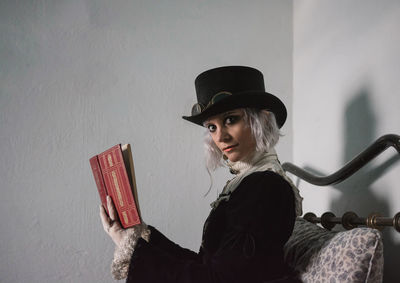 Portrait of young woman holding hat while sitting against wall