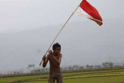 Shirtless boy holding flag while standing at farm against cloudy sky