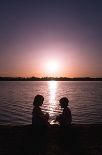 Silhouette boy sitting by lake against sky during sunset