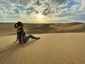 Mature woman sitting on sand dune in desert against cloudy sky