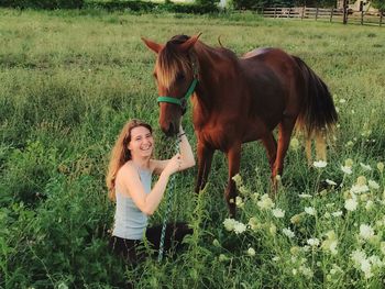 Portrait of cheerful young woman with brown horse on grassy field at farm