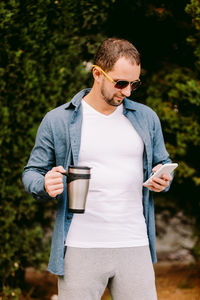 Man holding coffee cup while using mobile phone standing against trees
