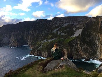 Woman sitting on cliff by sea against sky