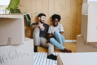 Happy multiracial couple sitting together against wooden wall at new home