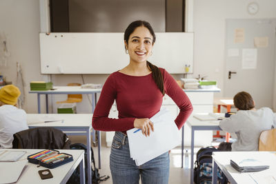 Portrait of smiling female teacher holding documents while standing in classroom