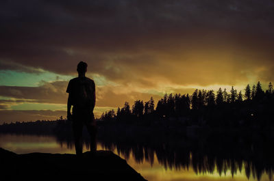 Rear view of silhouette man standing at lakeshore against sky during sunset