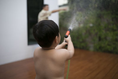 Rear view of shirtless boy spraying water at backyard