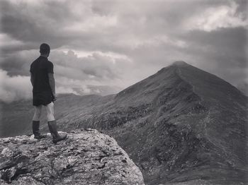 Rear view of man standing on cliff by mountains against cloudy sky