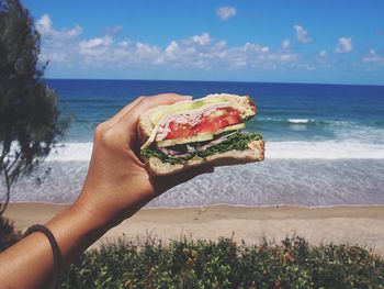 Cropped hand holding sandwich against sea