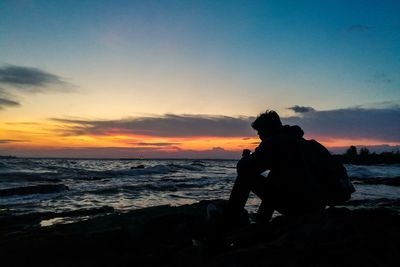 Silhouette man sitting on beach against sky during sunset