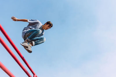 From below of brave male jumping over metal fence in street and showing parkour trick against blue sky