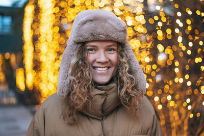 Portrait of young smiling woman with curly hair in fur hat in winter street decorated with lights