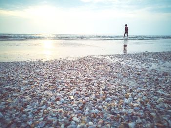 Man standing on shore at beach against sky during sunset