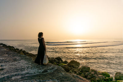 Rear view of woman standing at beach against sky during sunset