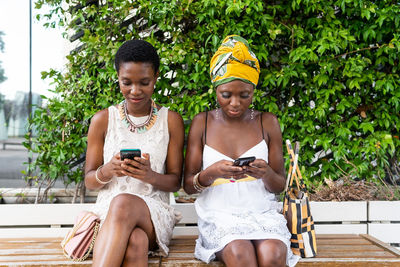 Adult african american female friends in stylish dresses and authentic accessories sitting on bench in city park and using smartphones