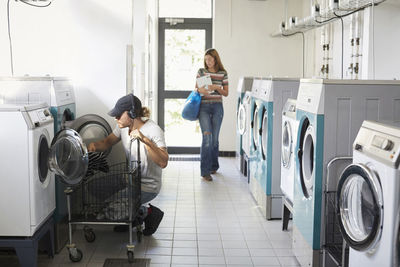 Man using washing machine while woman walking in laundromat