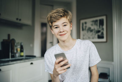 Portrait of smiling teenage boy holding smart phone while sitting in kitchen at home