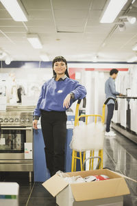 Portrait of smiling saleswoman standing by luggage cart in electronics store