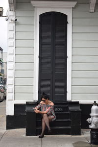 Woman using phone while sitting on steps against closed black door at sidewalk