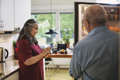 Side view of senior woman using digital tablet while man standing at refrigerator