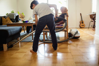 Boy cleaning floor with vacuum cleaner while mother sitting on armchair at home