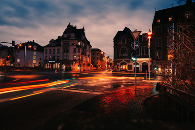 Light trails on road by buildings against sky at dusk