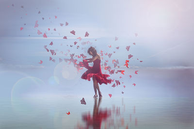 Digital composite image of woman wearing red dress dancing with butterflies on shore