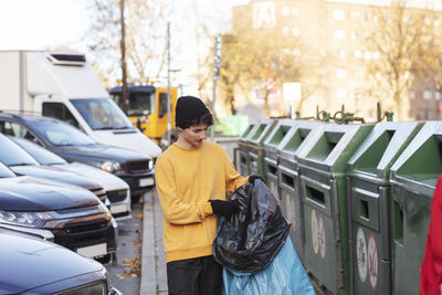 Young male volunteer holding waste in plastic bag by garbage can