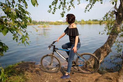 An adult woman with a bicycle stopped on the shore of a lake enjoying nature.